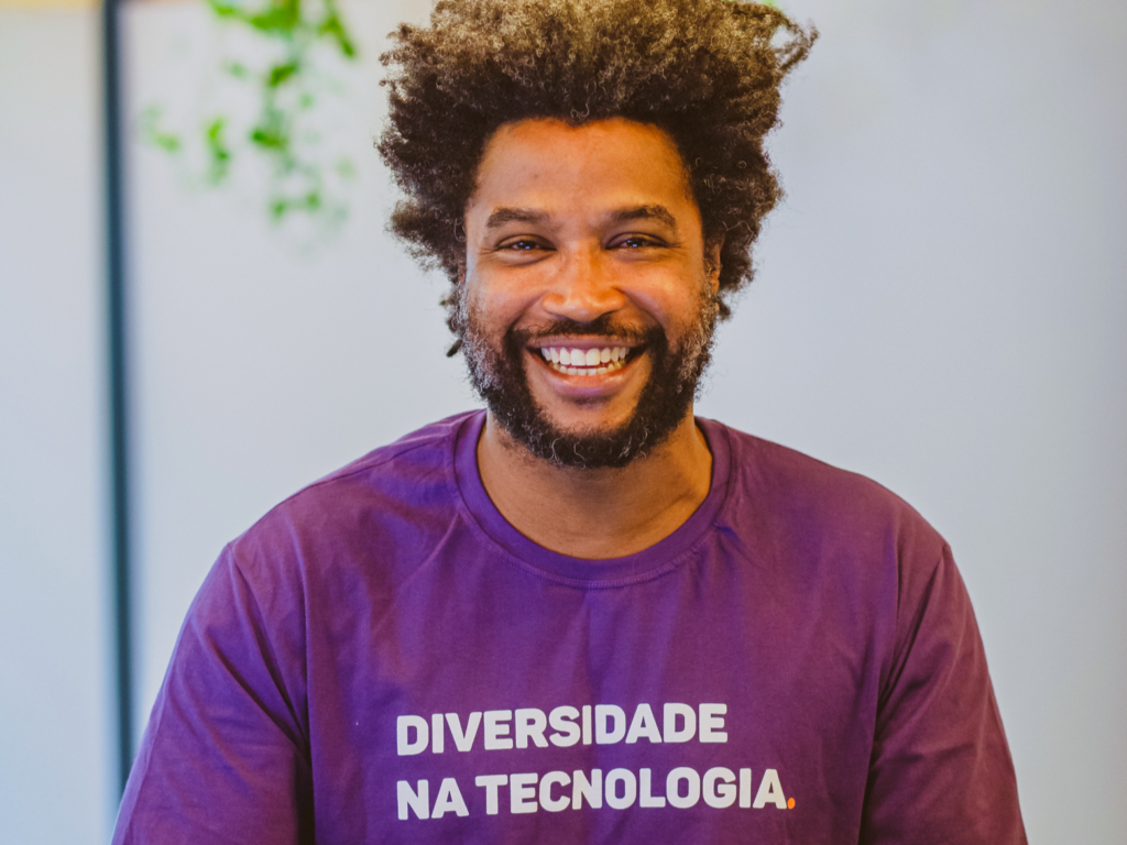 Na imagem, há um ex-aluno negro da Toti Diversidade vestindo uma camisa roxa escrito "Diversidade na Tecnologia". Ele está usando um notebook.