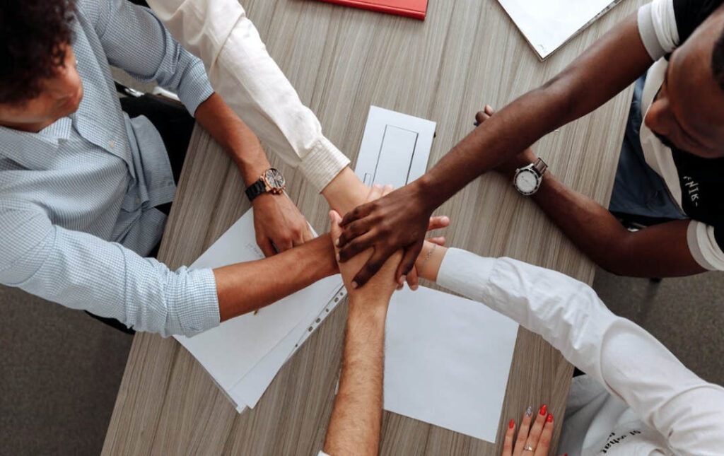 Na imagem, há pessoas colocando as mãos umas nas outras, remetendo a um gesto de trabalho em equipe. O fundo da foto lembra um ambiente corporativo.