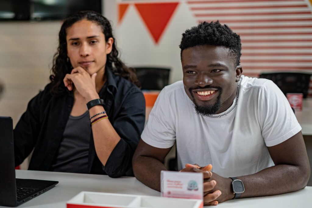 Duas pessoas do sexo masculino sentadas em uma mesa. Ambos foram estudantes da Toti Diversidade. Um deles é o Bryan, migrante da Colômbia. É indígena e tem cabelos na altura do ombro. O outro é o Seth, migrante de Gana, é negro e está sorrindo. 