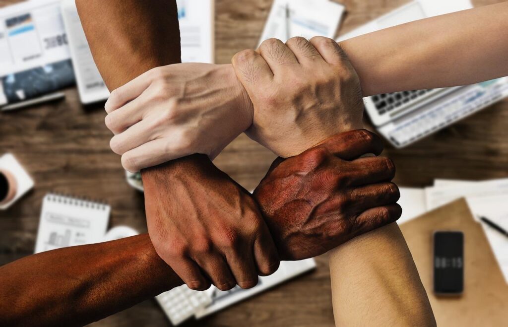 Na foto, há a imagem apenas de quatro braços de pessoas com raças diferentes, representando a diversidade e inclusão nas empresas. 